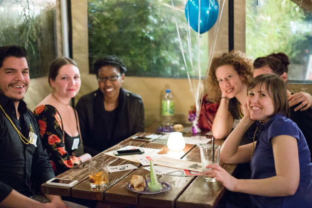 Group of six people enjoying a lively gathering at a table with drinks and snacks, celebrating the Love Bomb 2017 event supporting My Voice Music, with festive balloons and a warm atmosphere.